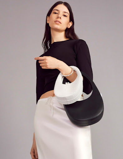 Fashion shot of a woman in a black top and white skirt holding two handbags, one black and one white, against a grey backdrop.