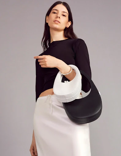 Fashion shot of a woman in a black top and white skirt holding two handbags, one black and one white, against a grey backdrop.