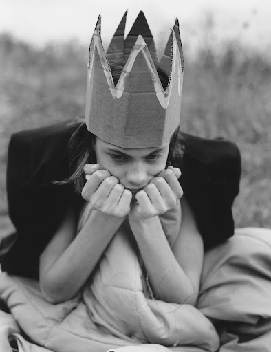 Monochromatic portrait of a child wearing a cardboard crown, sitting wrapped in a blanket in a field with a contemplative expression.