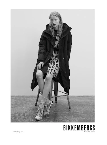 Black and white shot of a female fashion model in an overcoat and printed dress, seated on a stool against a plain backdrop.