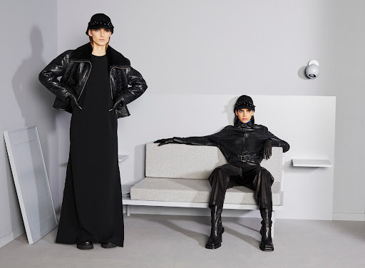 Two fashion models in black coats and hats pose inside a minimalist room with a grey sofa and walls.