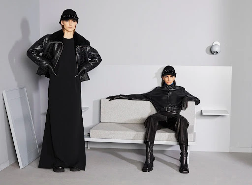 Two fashion models in black coats and hats pose inside a minimalist room with a grey sofa and walls.