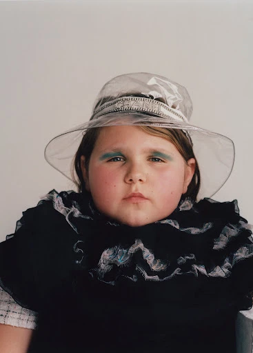 Young girl in a transparent hat and theatrical blue makeup posing in front of a white background, fashion editorial-style portrait.