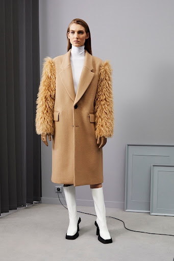 A woman in a tan coat with fur sleeves and white heeled boots poses in a minimalist indoor studio with grey walls and picture frames.