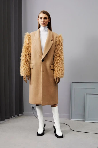 A woman in a tan coat with fur sleeves and white heeled boots poses in a minimalist indoor studio with grey walls and picture frames.