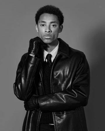 Black and white studio portrait of a young Black man wearing a leather jacket, tie, and leather gloves by Leon Mark