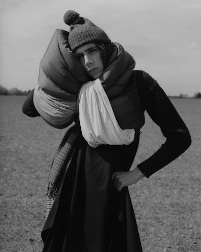 Monochrome portrait of a young adult wearing a knit hat and scarf, standing in a field under a bright sky, with dark tones.