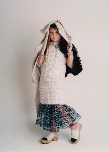 Child fashion model poses in tweed dress, jacket, jewelry, and wood clogs against a white background in a full studio shot.
