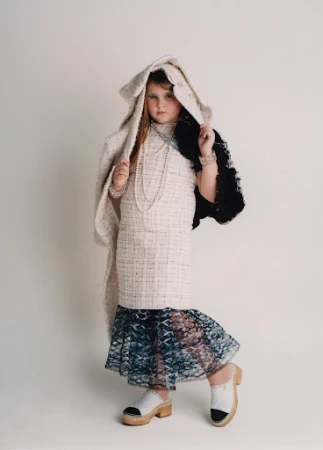 Child fashion model poses in tweed dress, jacket, jewelry, and wood clogs against a white background in a full studio shot.
