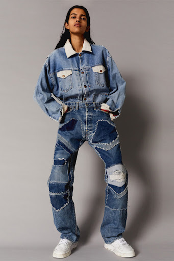 Full shot of a young adult in a light wash denim jacket with a white collar and patchwork denim pants and white sneakers against a grey backdrop.