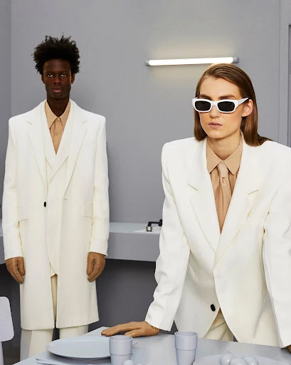 Two fashion models in white suits and beige shirts pose in a neutral-tone interior with minimal table props and an overhead light fixture.