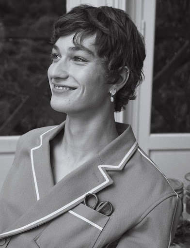 Black and white portrait of a smiling young adult with short curly hair, wearing pajamas and earrings indoors near a window.