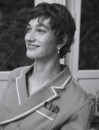Black and white portrait of a smiling young adult with short curly hair, wearing pajamas and earrings indoors near a window.