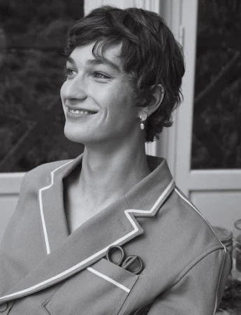 Black and white portrait of a smiling young adult with short curly hair, wearing pajamas and earrings indoors near a window.