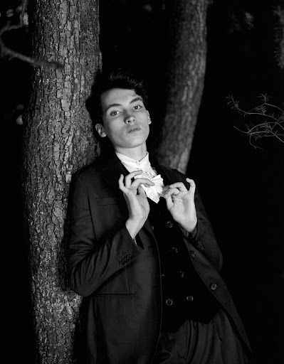 Monochrome portrait of a young adult male in a suit jacket and vest standing next to a tree in a dimly lit forest environment.