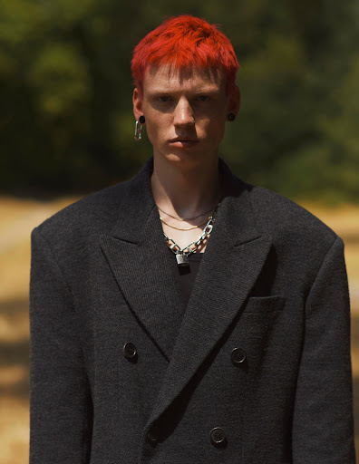 Portrait of a red-haired adult man wearing a dark grey wool double-breasted blazer, standing outdoors with a blurred background of trees.