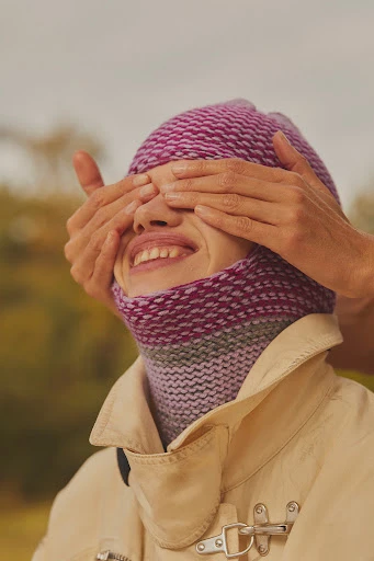 Smiling woman with hands covering her eyes, wearing a knitted hat and scarf, photographed outdoors with soft, diffused lighting.