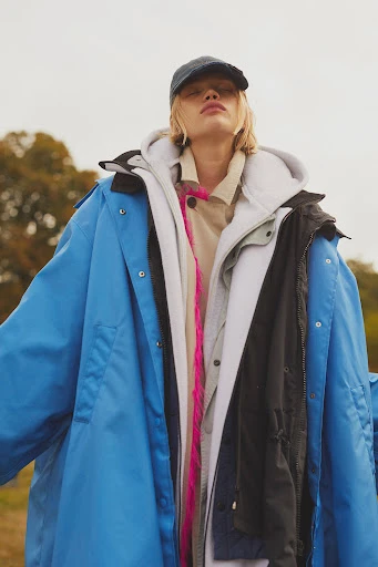 A person with blond hair wears a navy baseball cap and layers of jackets and hoodies in an outdoor setting on an overcast day.