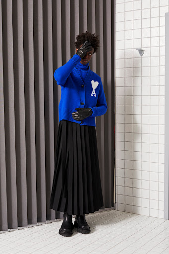 Adult male wearing a blue cardigan and a black pleated skirt posing in an indoor setting with gray vertical stripes and white tiles.