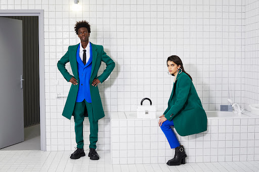 Two fashion models pose in colorful suits in a modern bathroom with white tile walls and floors; a handbag sits on the tub.