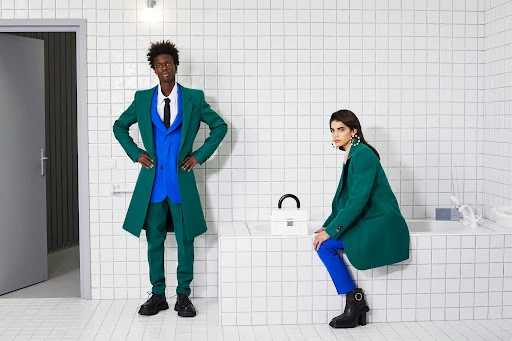 Two fashion models pose in colorful suits in a modern bathroom with white tile walls and floors; a handbag sits on the tub.