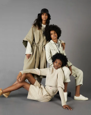 Three female models posing together against a plain grey studio backdrop wearing neutral-toned apparel and footwear.
