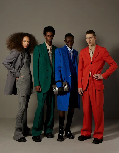 Four fashion models wearing color block suits posing indoors against a grey backdrop in a fashion editorial photography studio shot.