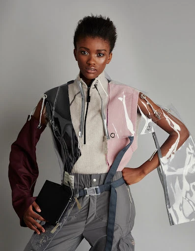 Fashion model in clear vinyl top, grey pants, and black bag poses in a photo studio with a grey background for a fashion editorial shoot.