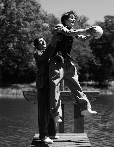 Black and white photograph of two adult males on a wooden dock, one lifting the other who is holding a soccer ball outdoors.