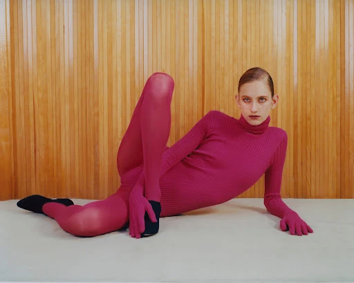 A fashion portrait features a reclining, fair-skinned young woman in a pink ribbed turtleneck bodysuit against a simple wood panel backdrop.