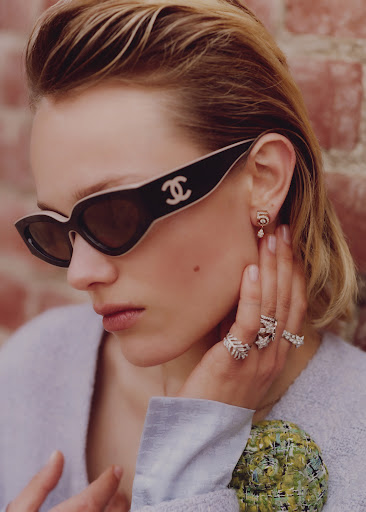 A woman poses, wearing Chanel sunglasses, an earring, and rings, with a light purple top and a brick wall in the background.
