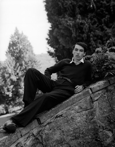 Monochrome portrait of a young man reclining on a stone wall outdoors against a backdrop of trees