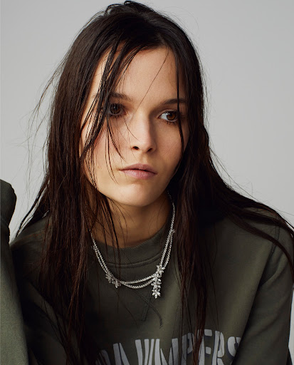 Close-up of a young woman with dark hair wearing a green sweatshirt and necklace, against a grey background, fashionable look.