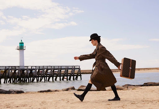 Man in coat and hat walking on beach carrying briefcase in a fashion editorial, with lighthouse and dock in the background.