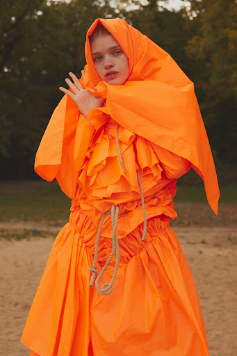 Model in vibrant orange hooded dress with ruffled details and rope accents outdoors by Leon Mark