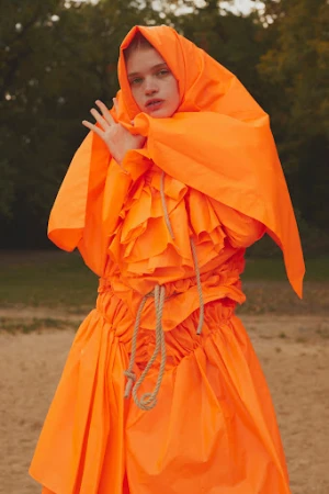 Model in vibrant orange hooded dress with ruffled details and rope accents outdoors by Leon Mark