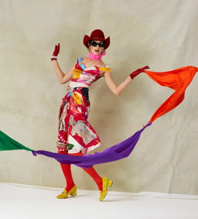 Model in a red cowboy hat and patterned dress holding flowing colorful scarfs by director Leon Mark