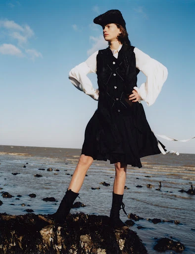 A young adult stands on a rocky beach, wearing a black vest, skirt, and hat, paired with a white blouse, against a blue sky and horizon.