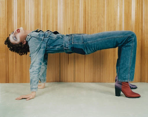 Woman in denim jumpsuit and heels strikes an arch pose against a wood paneled wall and light carpet in a fashion editorial shot.