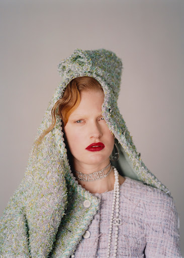Woman with red hair and red lipstick wears a light purple tweed jacket with Chanel jewelry and a hood in a studio setting.