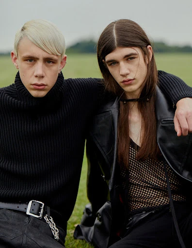 Two young men pose outdoors, one with platinum blonde hair and a black turtleneck, the other with long brown hair and a black leather jacket.