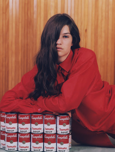 Brunette woman in a red shirt poses with a stack of Campbell's Tomato Soup cans against a wood panel background in a studio setting.