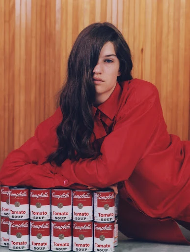 Brunette woman in a red shirt poses with a stack of Campbell's Tomato Soup cans against a wood panel background in a studio setting.
