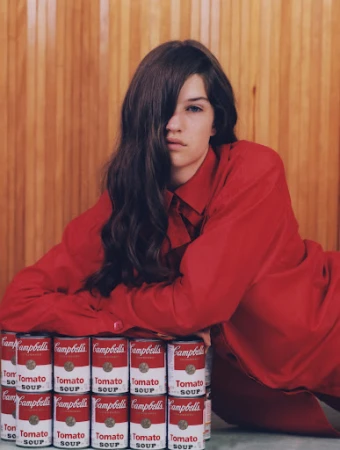 Brunette woman in a red shirt poses with a stack of Campbell's Tomato Soup cans against a wood panel background in a studio setting.