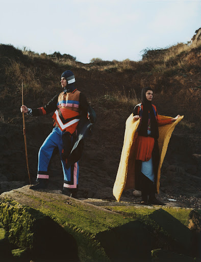 Two people in costume standing on a rocky outcrop with coastal vegetation under a bright sky, looking off into the distance.