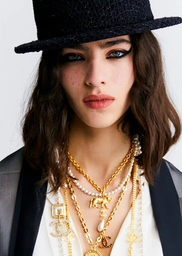 Close-up of woman wearing a hat and multiple strands of gold necklaces. Heavily lined eyes and modern fashion style.