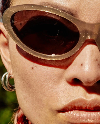 Close-up of an adult female wearing sunglasses and hoop earrings, with partial view of her face and a blurred green background.