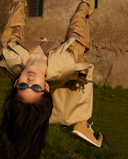 Model hanging upside down wearing sunglasses and a camel trench coat outdoors against a stone building
