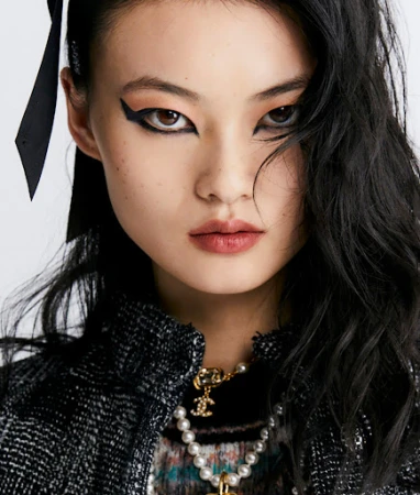 Close-up portrait of an East Asian woman with dramatic black winged eyeliner, wearing a dark tweed jacket and pearl necklace.