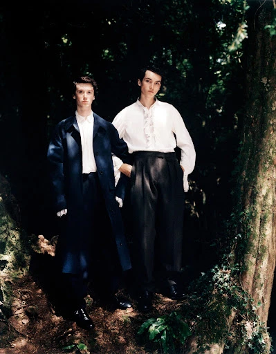 Two young adult men pose in a sun-dappled forest setting, wearing dress pants, collared shirts, and blazers.